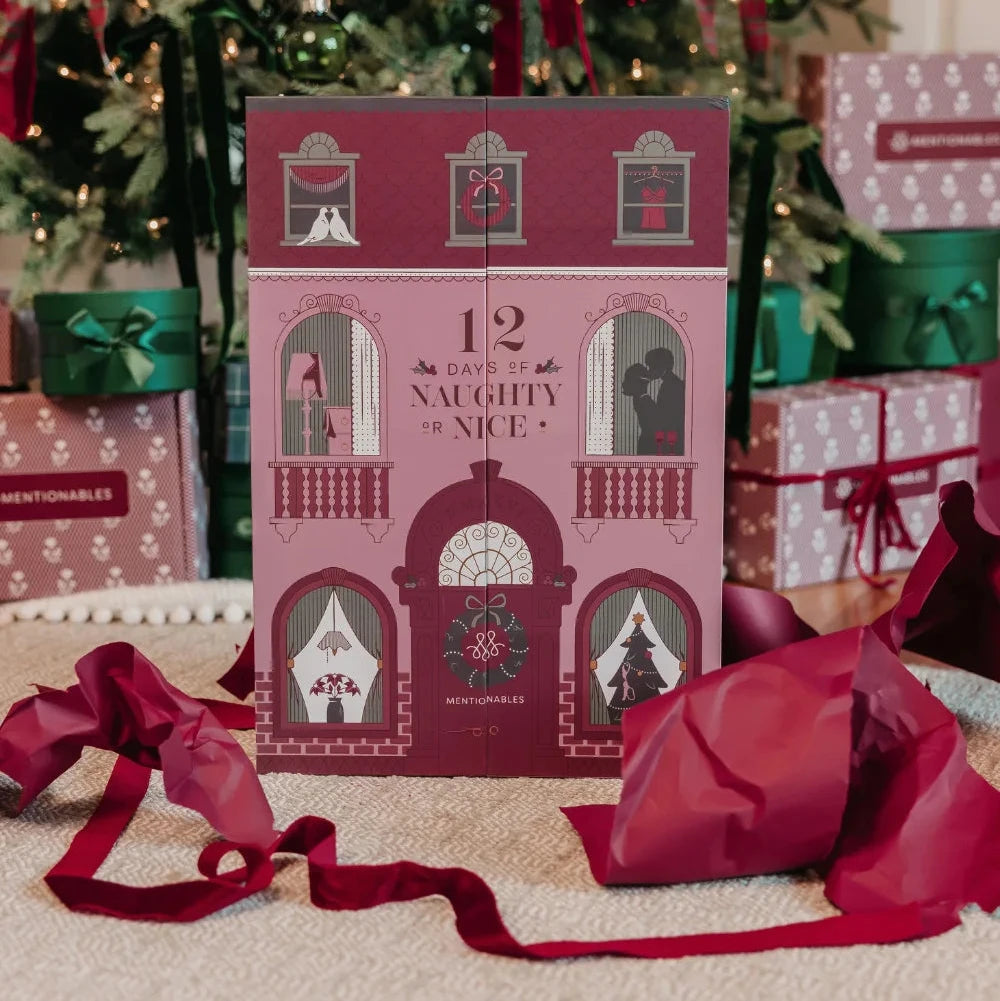 Mentionables Lingerie Christmas advent calendar card featuring a pink house illustration with the text '12 Days of Naughty or Nice,' styled on a carpet in front of a decorated Christmas tree with wrapped gifts and red ribbons.