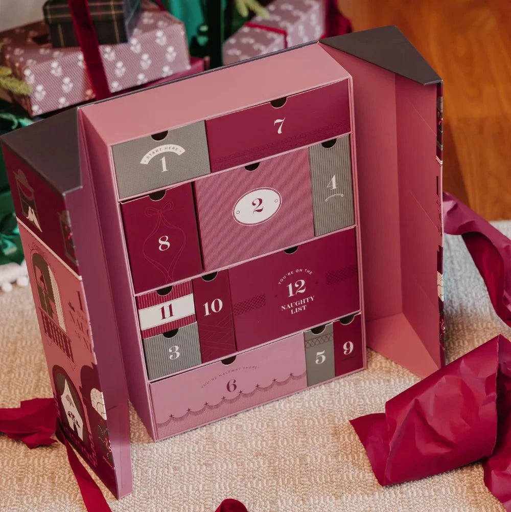 Mentionables Lingerie Christmas advent calendar card featuring a pink house illustration with the text '12 Days of Naughty or Nice,' styled on a carpet in front of a decorated Christmas tree with wrapped gifts and red ribbons. Open showing the reusable drawers in pink, cranberry, and green.