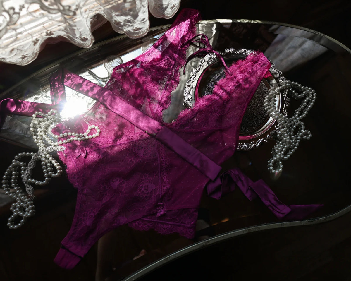A plum purple lace lingerie set with a matching belt and bow, displayed on a reflective dark surface with decorative pearl strands and a mirror.