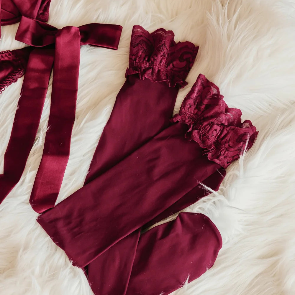Lacy Thigh High Stockings - Cranberry. A pair of burgundy thigh high tights with matching ruffled lace cuffs. Close up of the lace laid out on a white faux fur blanket.